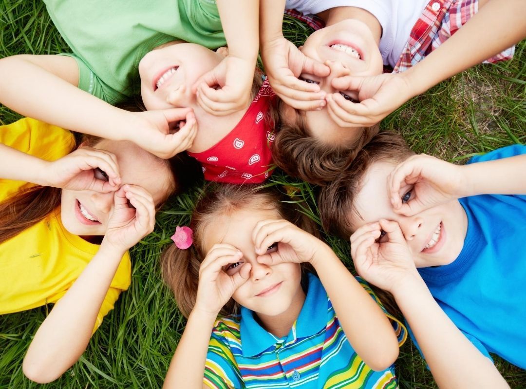 Children lying in a circle on grass, smiling and making eye shapes with their hands