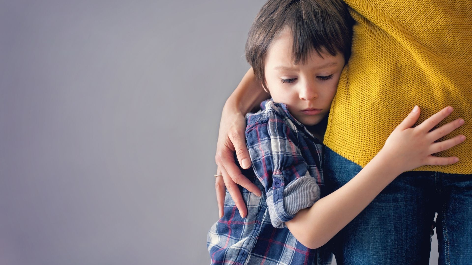 Child hugging an adult’s waist, with an arm around them, against a plain gray background.