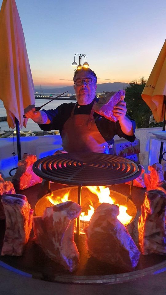 Chef che griglia la carne su fiamme libere. Scenario all'aperto con il tramonto sullo sfondo.