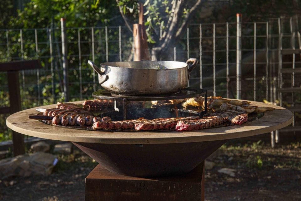Griglia da esterno con superficie di cottura circolare e spiedini di carne in cottura. Sopra è appoggiata una pentola, da cui fuoriesce il fumo.