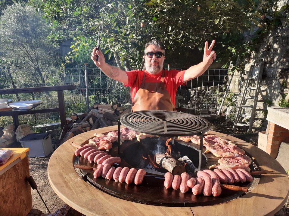 Uomo che griglia carne all'aperto, alzando le braccia per l'eccitazione. Salsicce e bistecche su una grande griglia circolare.