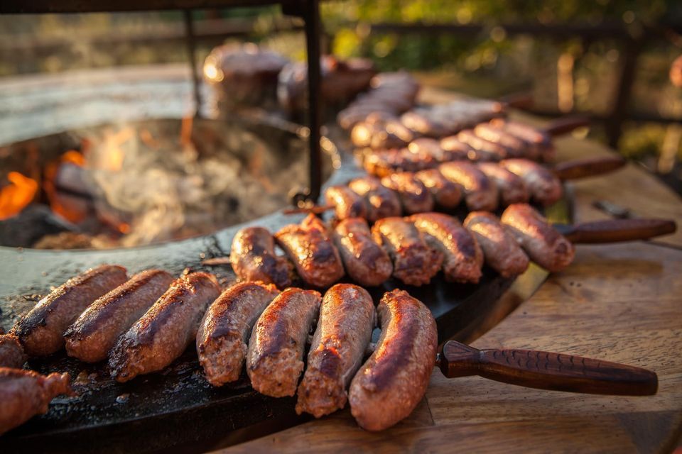 Salsicce cotte alla griglia su fuoco aperto, spiedini di legno, ambiente rustico.