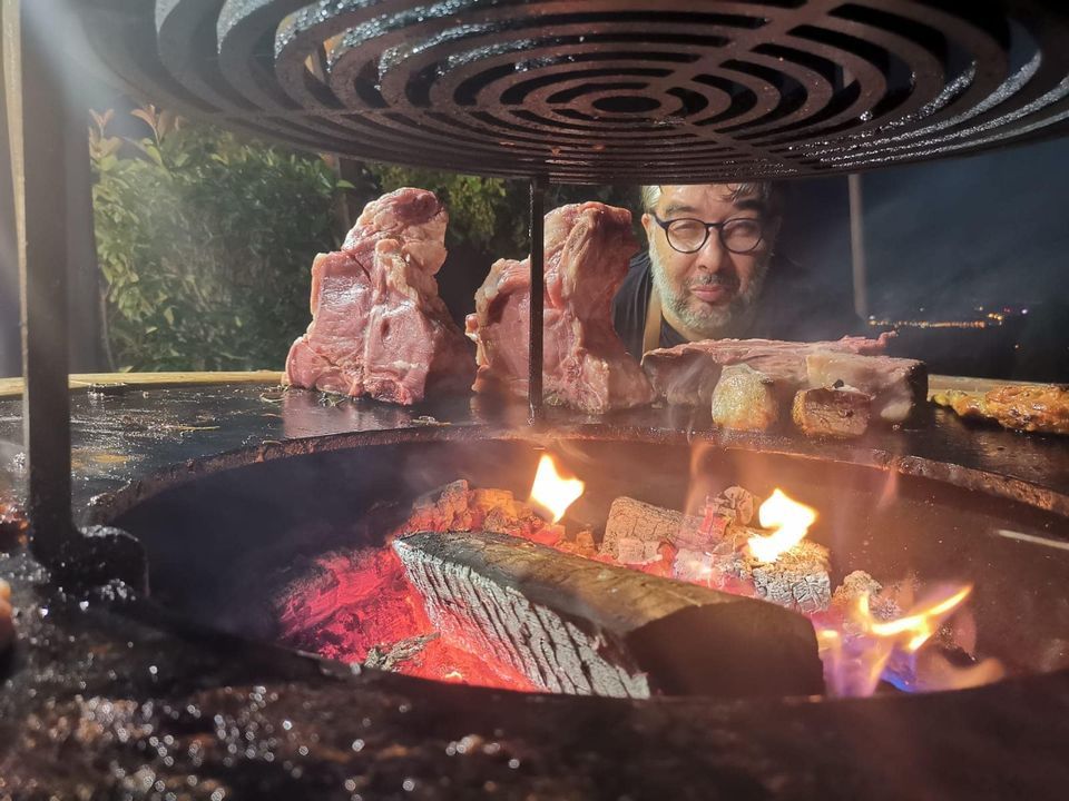 Uomo che griglia bistecche su un fuoco di legna. Bistecche sulla griglia; uomo che sbircia in basso, fiamme illuminate.
