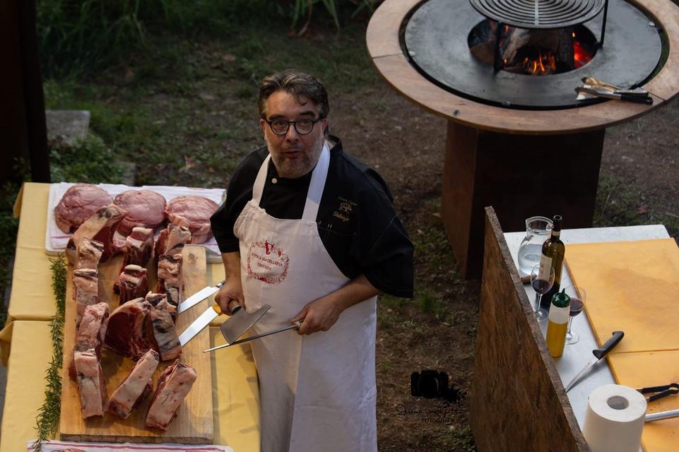 Uno chef con tagli di carne su una tavola di legno, vicino a una griglia con fuoco.