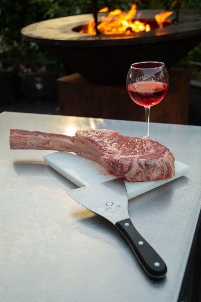 Bistecca tomahawk, bicchiere di vino e spatola su un tavolo in acciaio inossidabile con braciere sullo sfondo.
