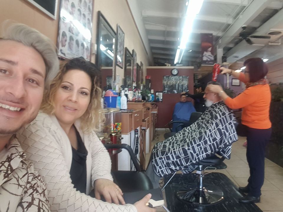 People in a barbershop. Two smiling people take a selfie. A barber blow-dries a customer's hair.