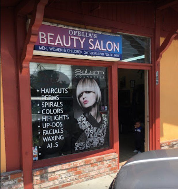 Ofelia's Beauty Salon storefront, red trim, sign for haircuts, perms, colors, and more. A person is seen inside.