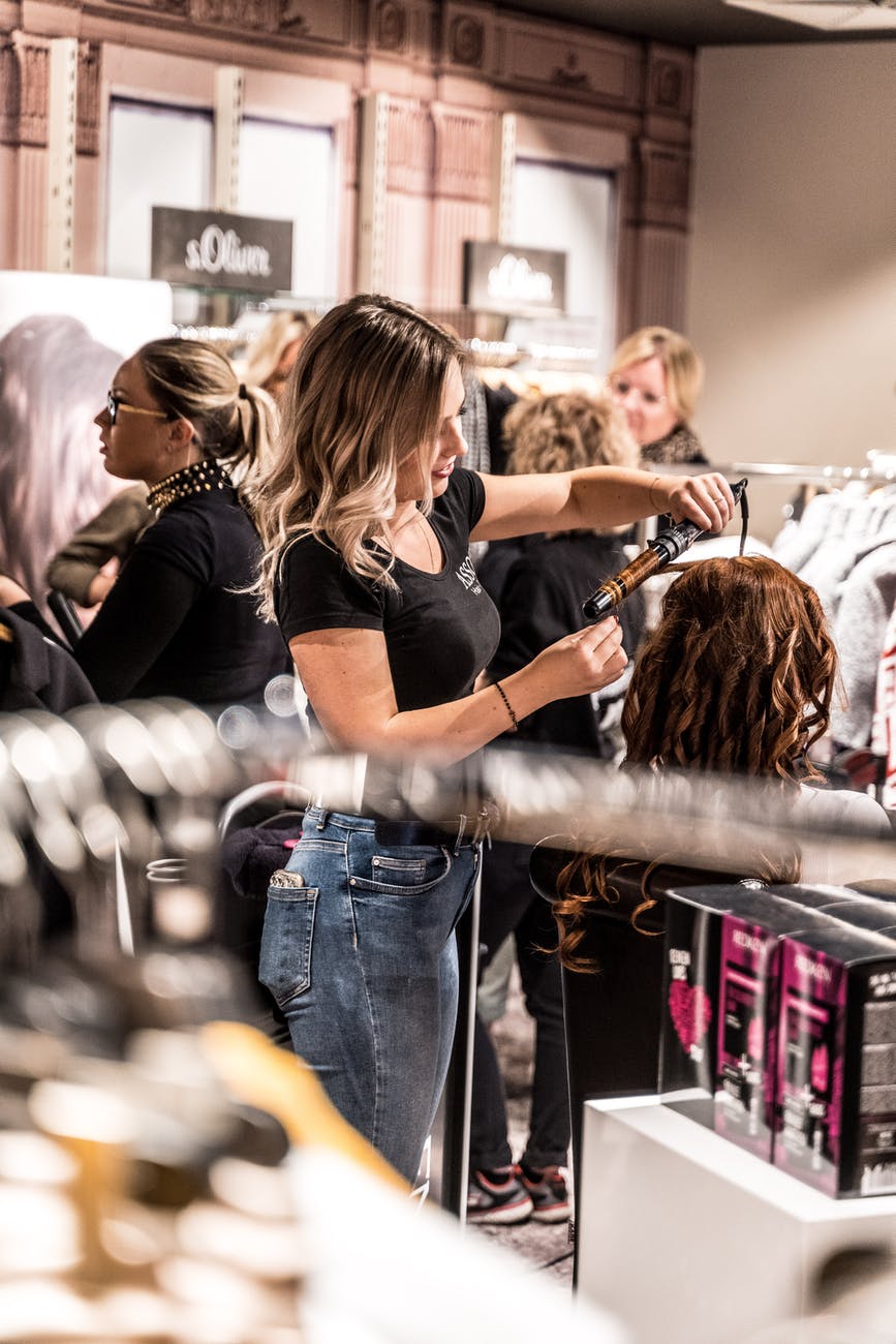 Woman styling hair on a mannequin in a clothing store. Other shoppers and staff are visible.