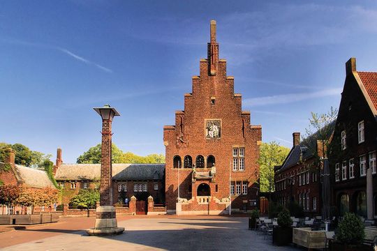 Op een stadsplein staat een historisch gebouw van rode baksteen met een trapgevel, naast een hoge, decoratieve stenen pilaar.