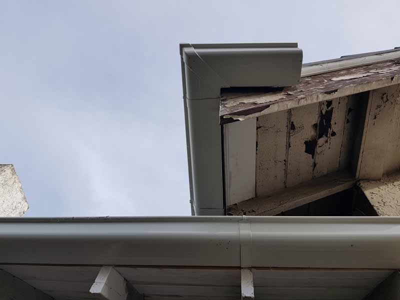 A low-angle view of a grey metal roof gutter system attached to the weathered, peeling wooden eaves of a house.