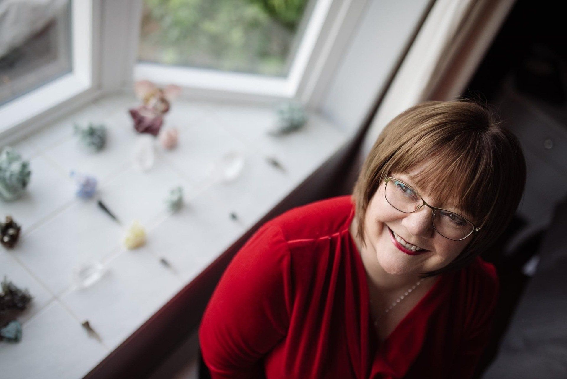 Smiling woman in a red top by a bright window with small figurines on the sill