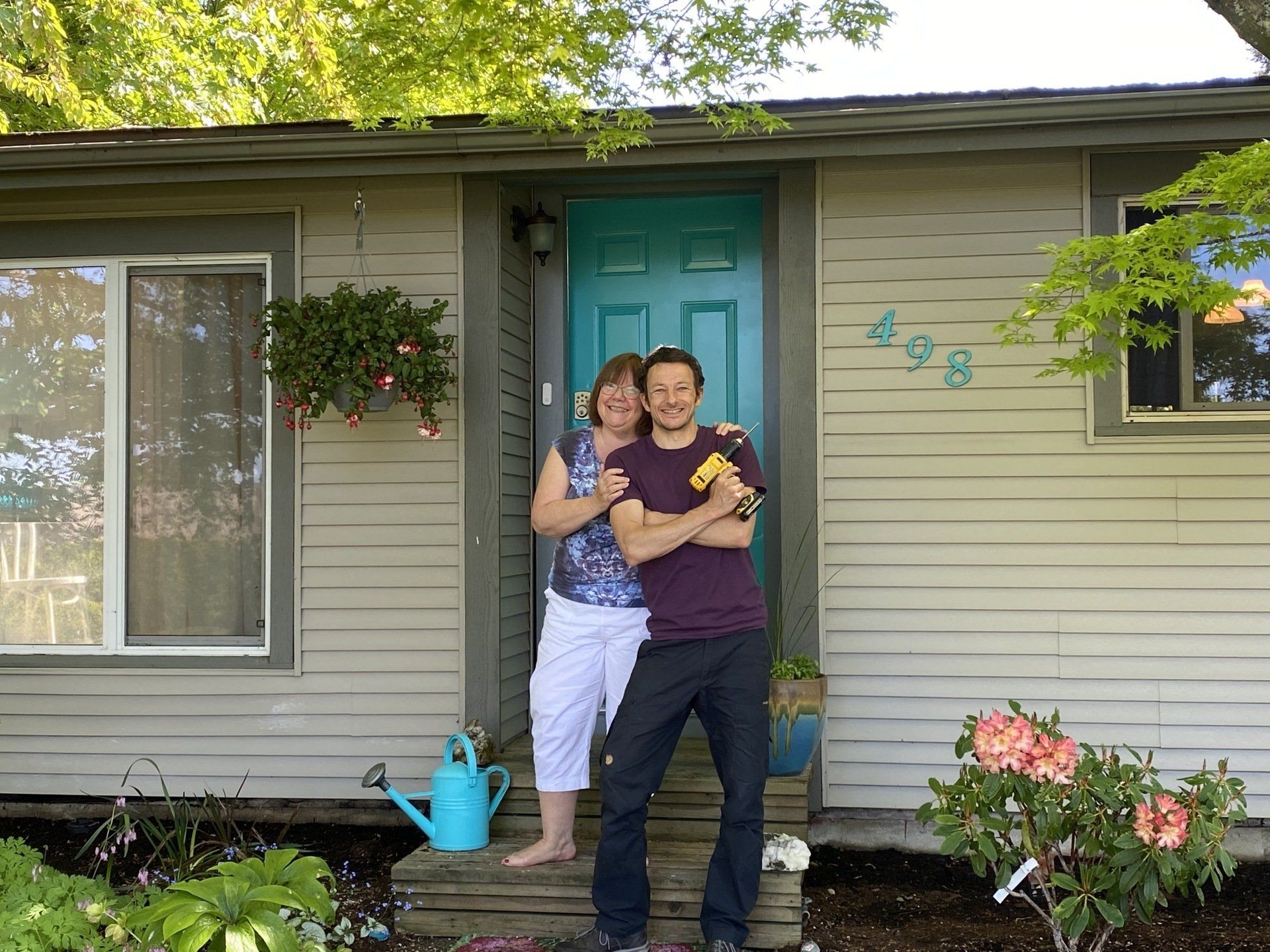 Two people standing and smiling in front of a teal front door, with hanging flowers and potted plants on the porch.