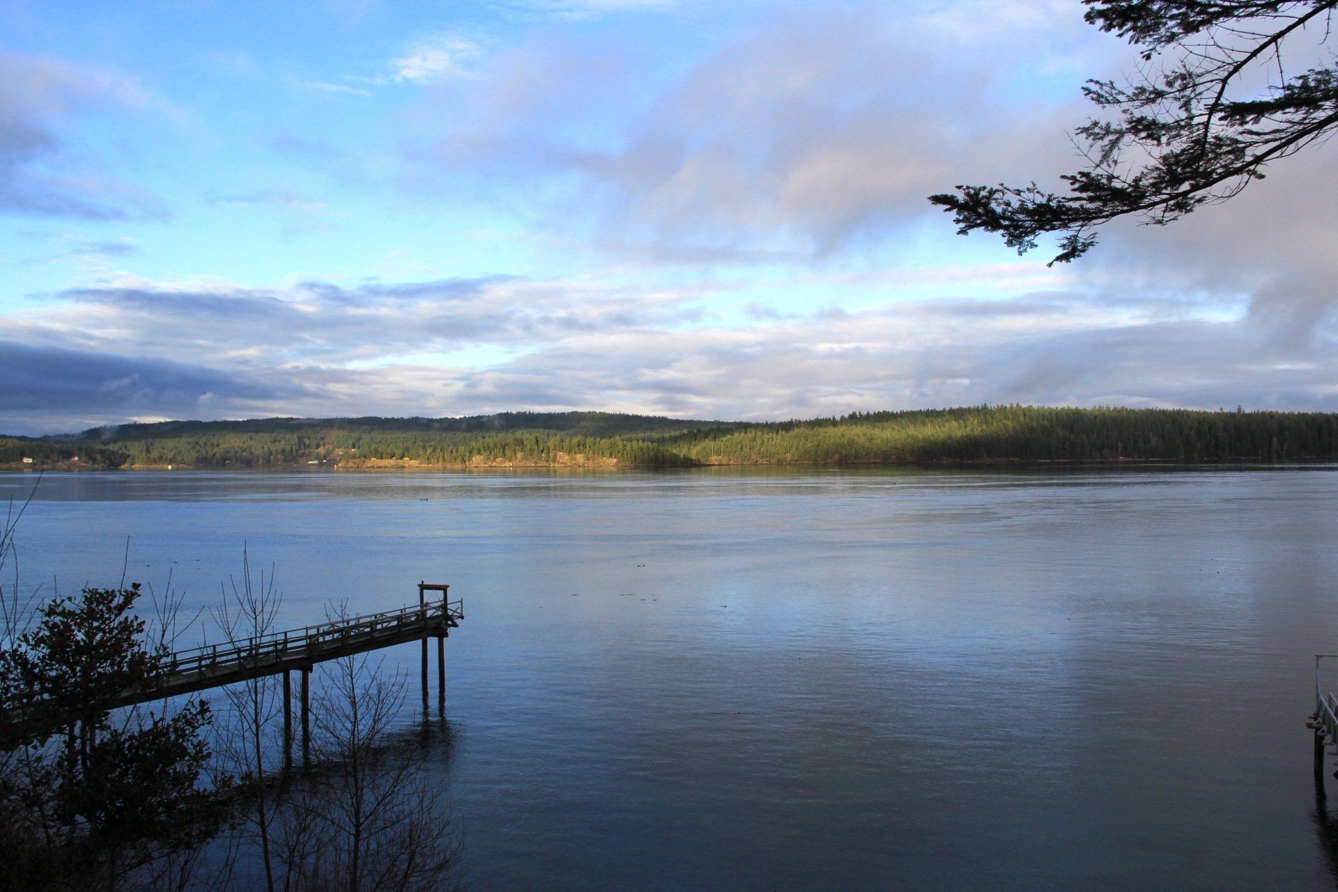 Calm lake under a cloudy blue sky, with a distant shoreline and a small dock in the foreground