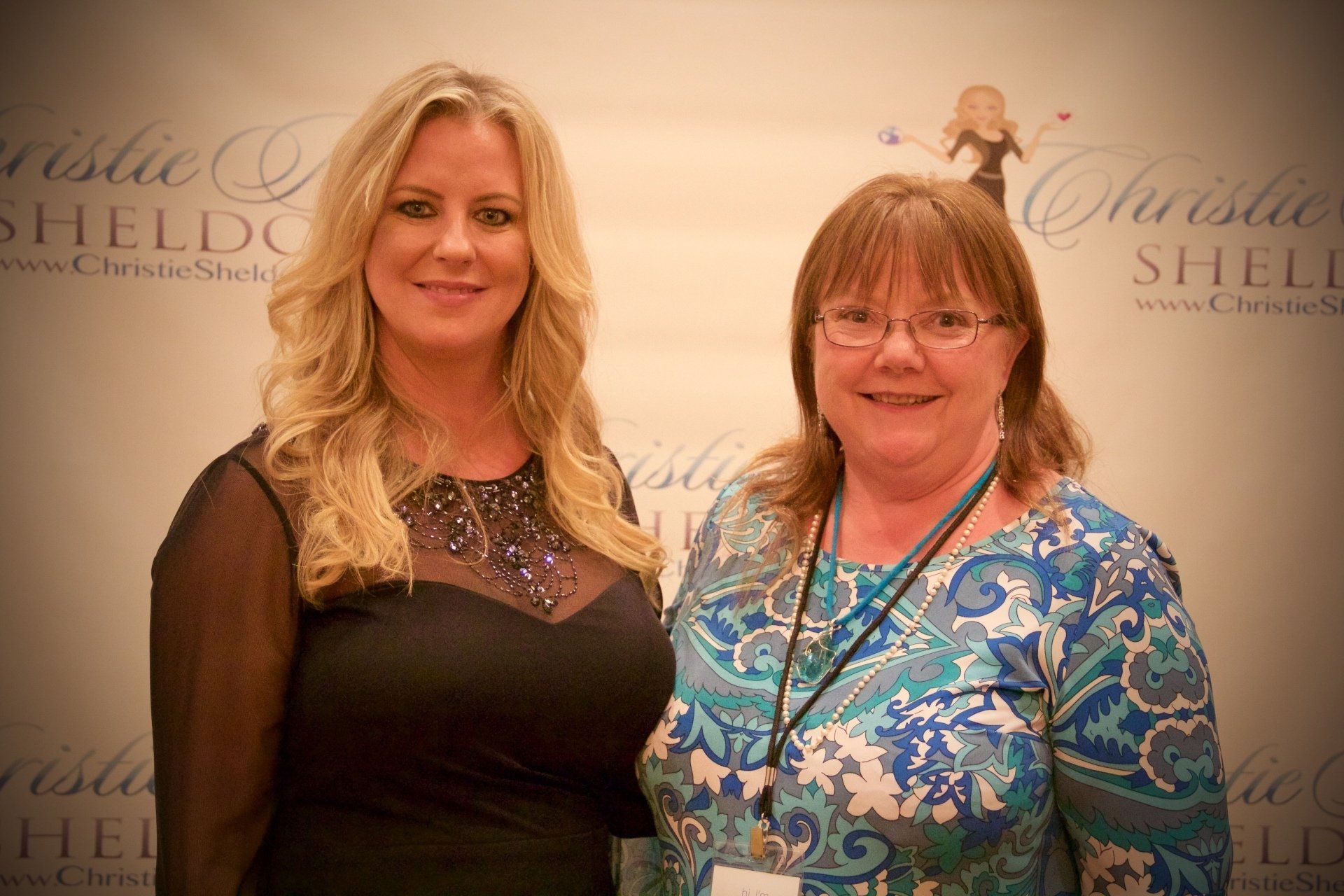 Two women posing at a step-and-repeat backdrop, wearing black and blue patterned dresses