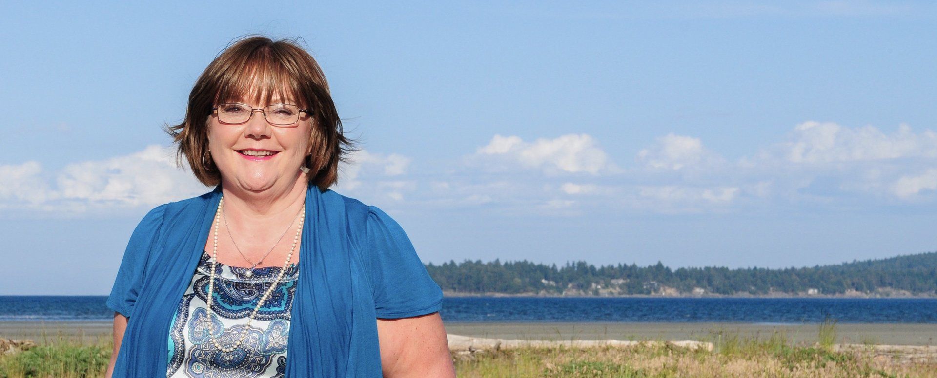 Smiling person in a blue cardigan standing outdoors by a lake under a clear sky