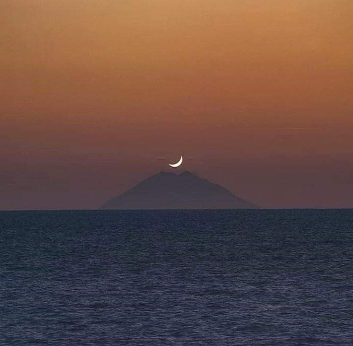 Una luna crescente sorge sull'oceano con una montagna sullo sfondo.
