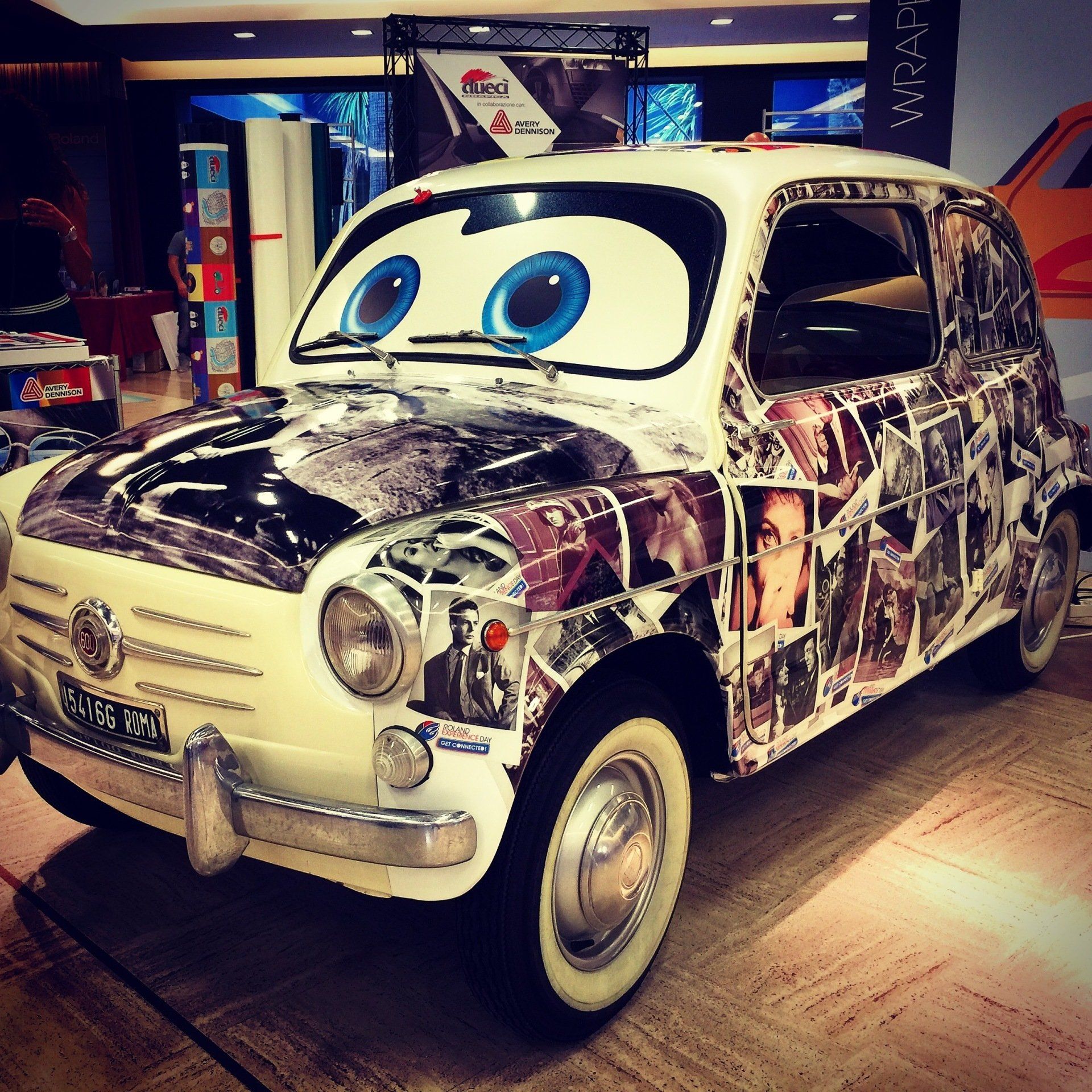 White Fiat 600 car decorated with photo collage and cartoon eyes. Inside a building.