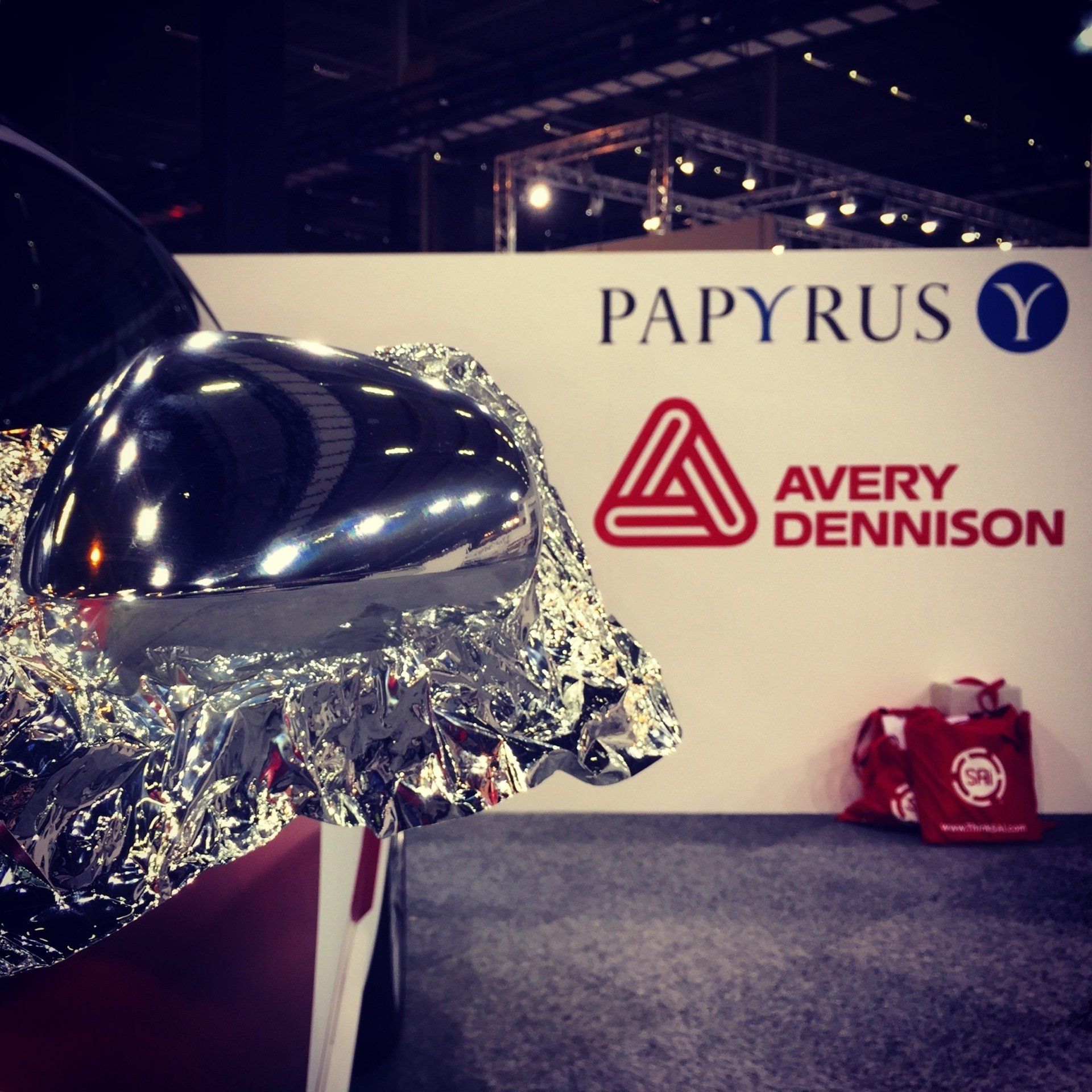 Shiny car mirror wrapped in foil, in front of a PAPYRUS and Avery Dennison display.