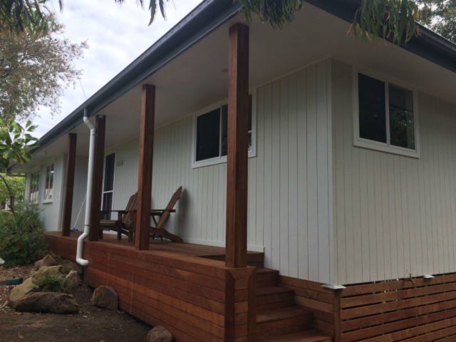 Single-story white house with a covered porch, wooden steps, and raised deck on a wooded lot