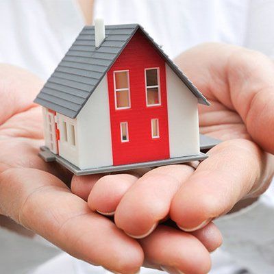 Hands cradling a miniature house model, white and red facade, grey roof, symbolizing home ownership.