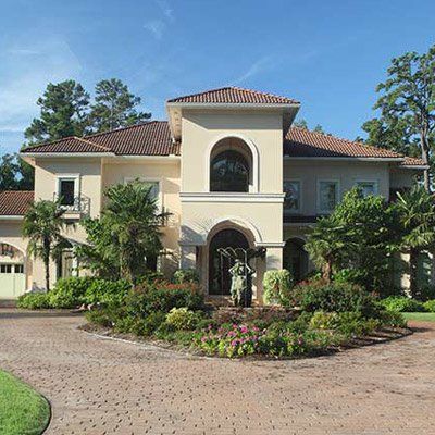 Beige stucco house with red tile roof, arched entry, and lush landscaping on a brick driveway.
