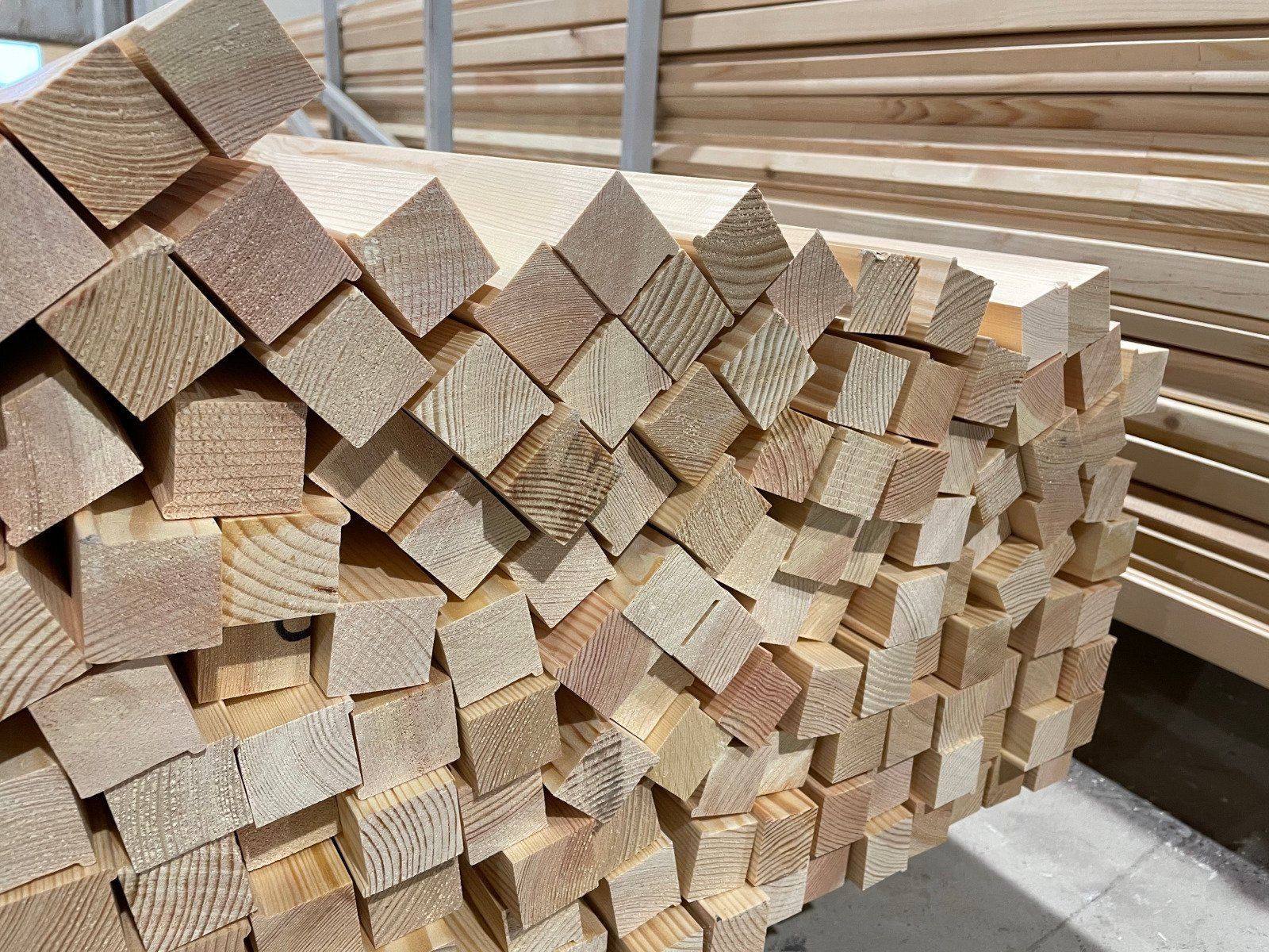 Stacked wooden lumber beams in a warehouse, showing cut ends of pale wood pieces.
