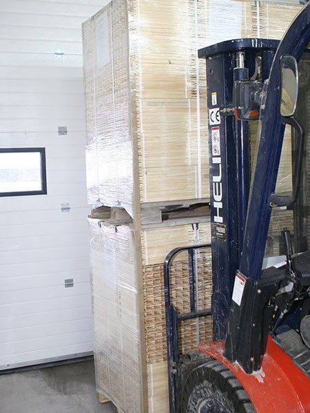Forklift lifting a wrapped pallet in a warehouse, beside a white wall and window.