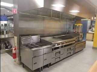 Stainless steel commercial kitchen with griddle, stove, and warming drawers, against a metal backsplash.