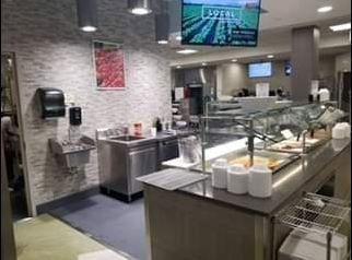 Food service area with stainless steel counter, sink, and buffet line; TV screen above.