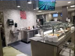 A cafeteria area with stainless steel serving counters, a hand-washing station, and a television displaying 