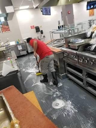 Person mopping a commercial kitchen floor with cleaning solution; stainless steel equipment, bright overhead lights.
