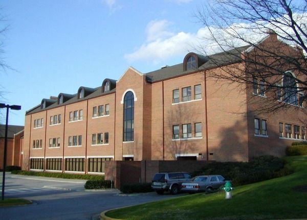 Red brick building with multiple windows, cars in parking area, grassy hill.