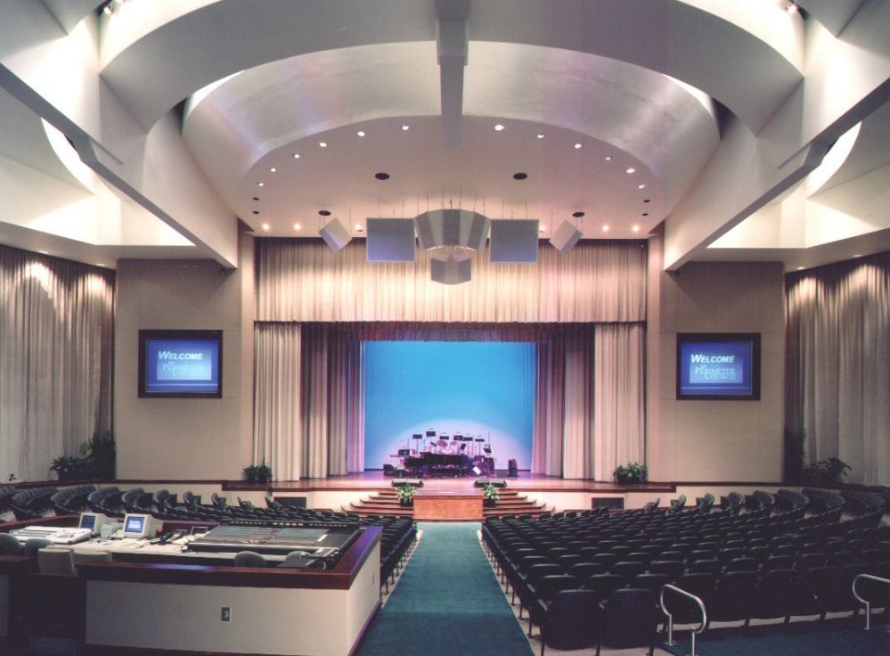 Auditorium with stage, curtains, blue backdrop, and rows of seats. Two screens display text.