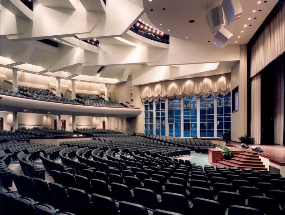 Interior of a large auditorium with rows of seats facing a stage with a podium. Large windows.