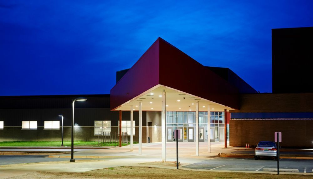 Red-roofed building entrance at night. Beige walkway, streetlights, blue sky.