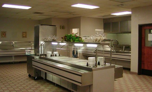 Empty cafeteria with stainless steel serving counters, beige walls, and a red door.