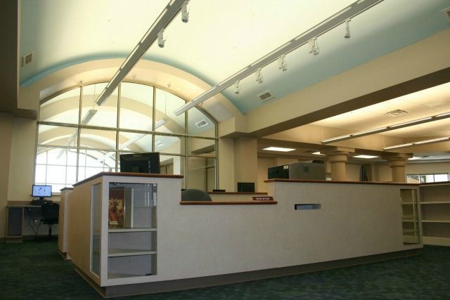 Library reception desk with computer screens, green carpet, and arched window in the background.