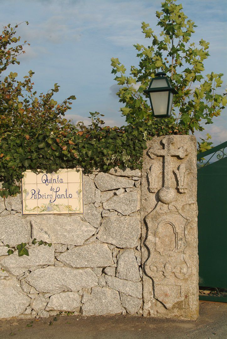 Stone wall with lantern, sign, and carved emblem. Lush greenery and gate in background.