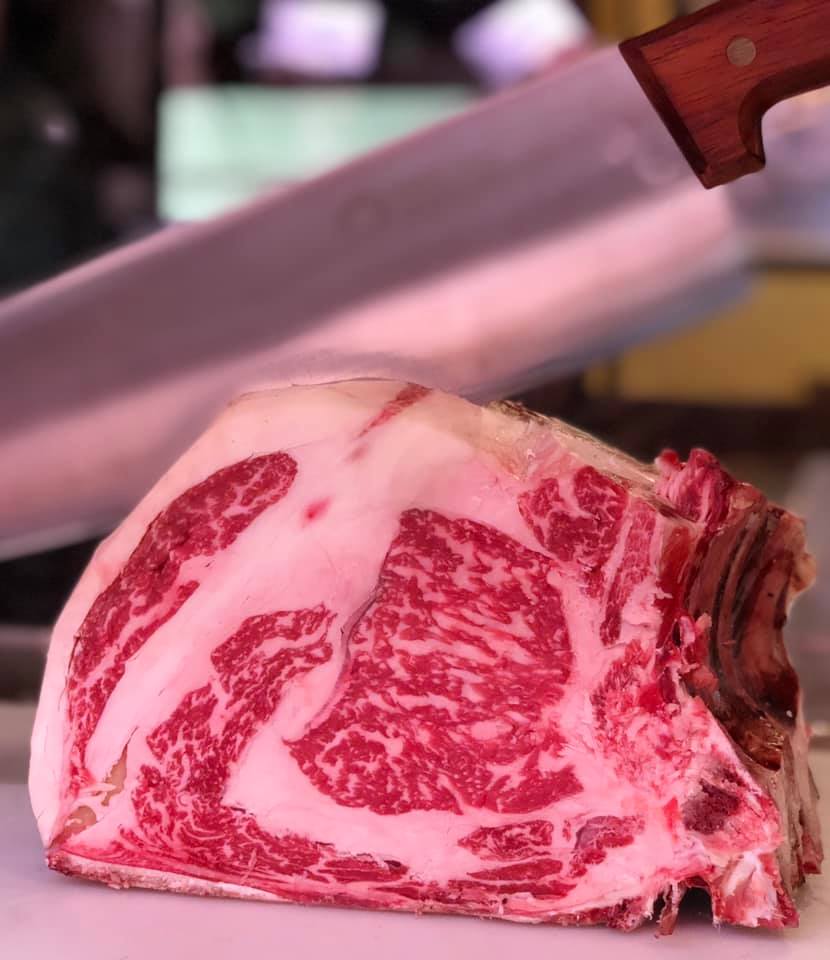 Primo piano di una grande bistecca di ribeye cruda con venature di grasso, con un coltello da macellaio sullo sfondo.