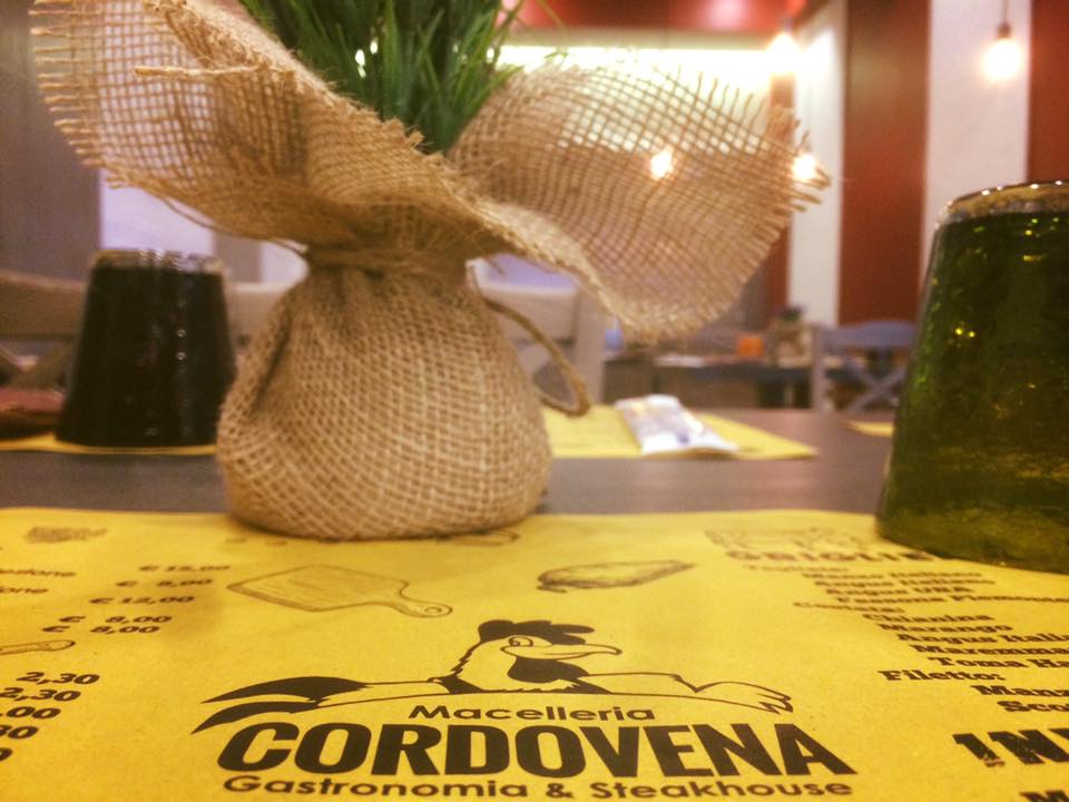 Apparecchiatura del tavolo al Cordovena Gastronomia & Steakhouse: pianta avvolta in tela di iuta, tovaglietta gialla con logo, bicchiere verde.