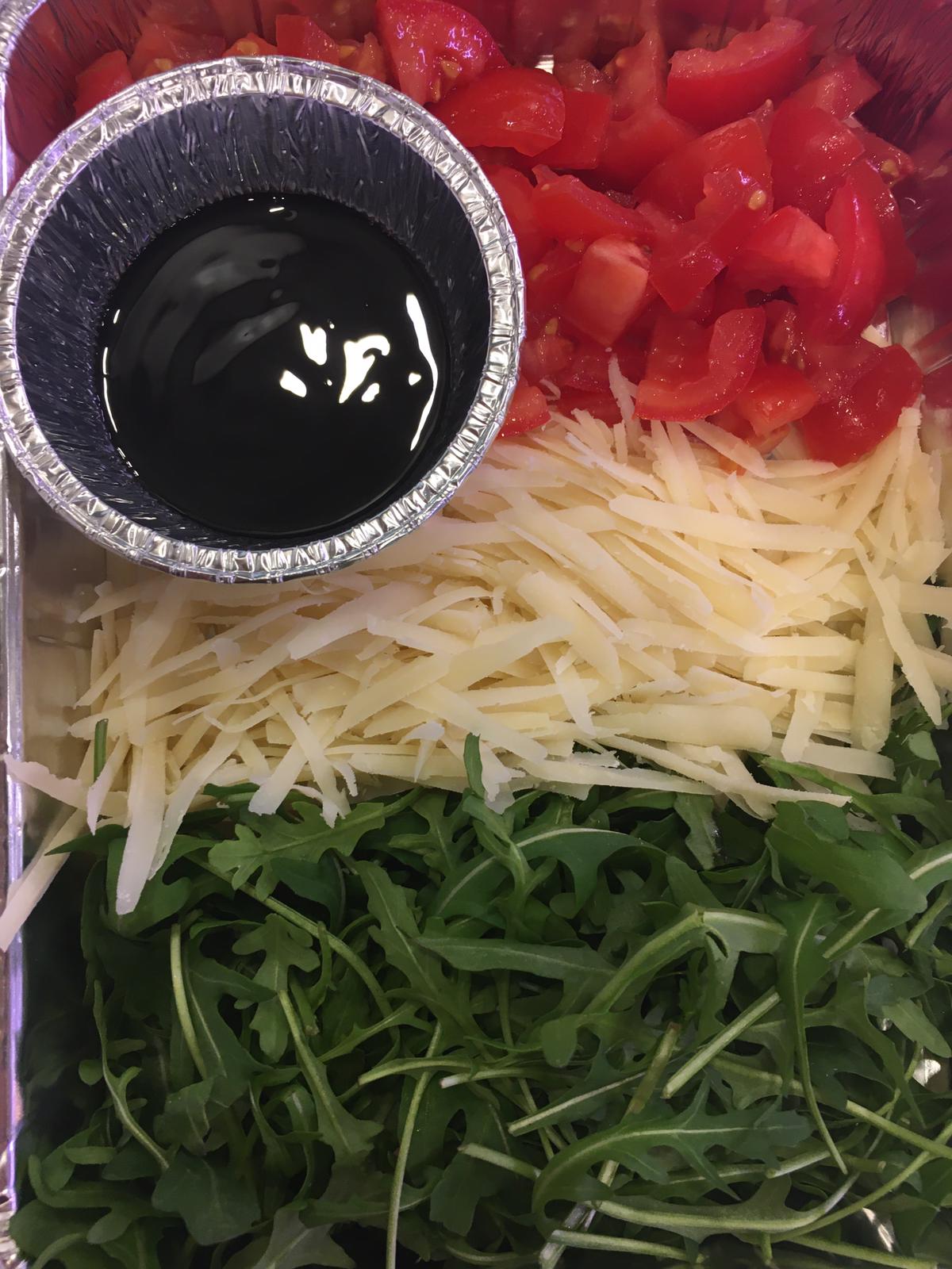 Un vassoio con gli ingredienti: pomodori, formaggio grattugiato, rucola e aceto balsamico.