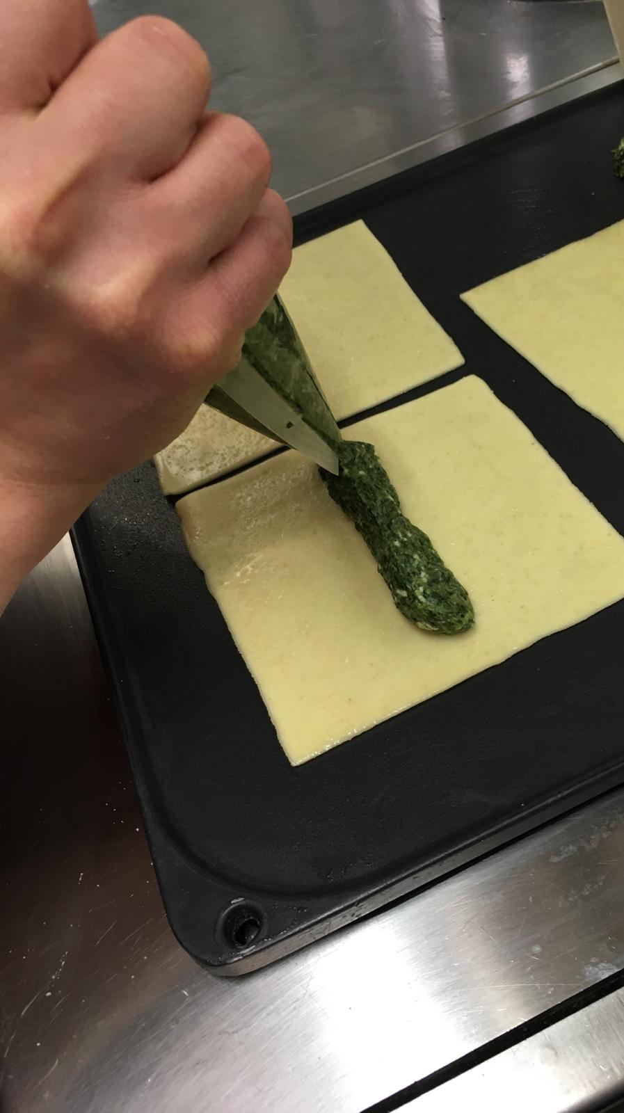 Una mano che spreme il ripieno verde sui quadrati di pasta usando una sac à poche su una superficie nera.