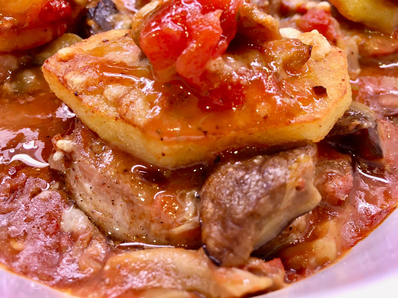 Fette di patate e carne cotte in una ricca salsa rossa con funghi e pomodori ben visibili.