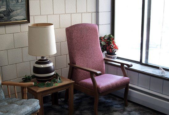 Chair with pink upholstery next to a small table with a lamp, by a window.