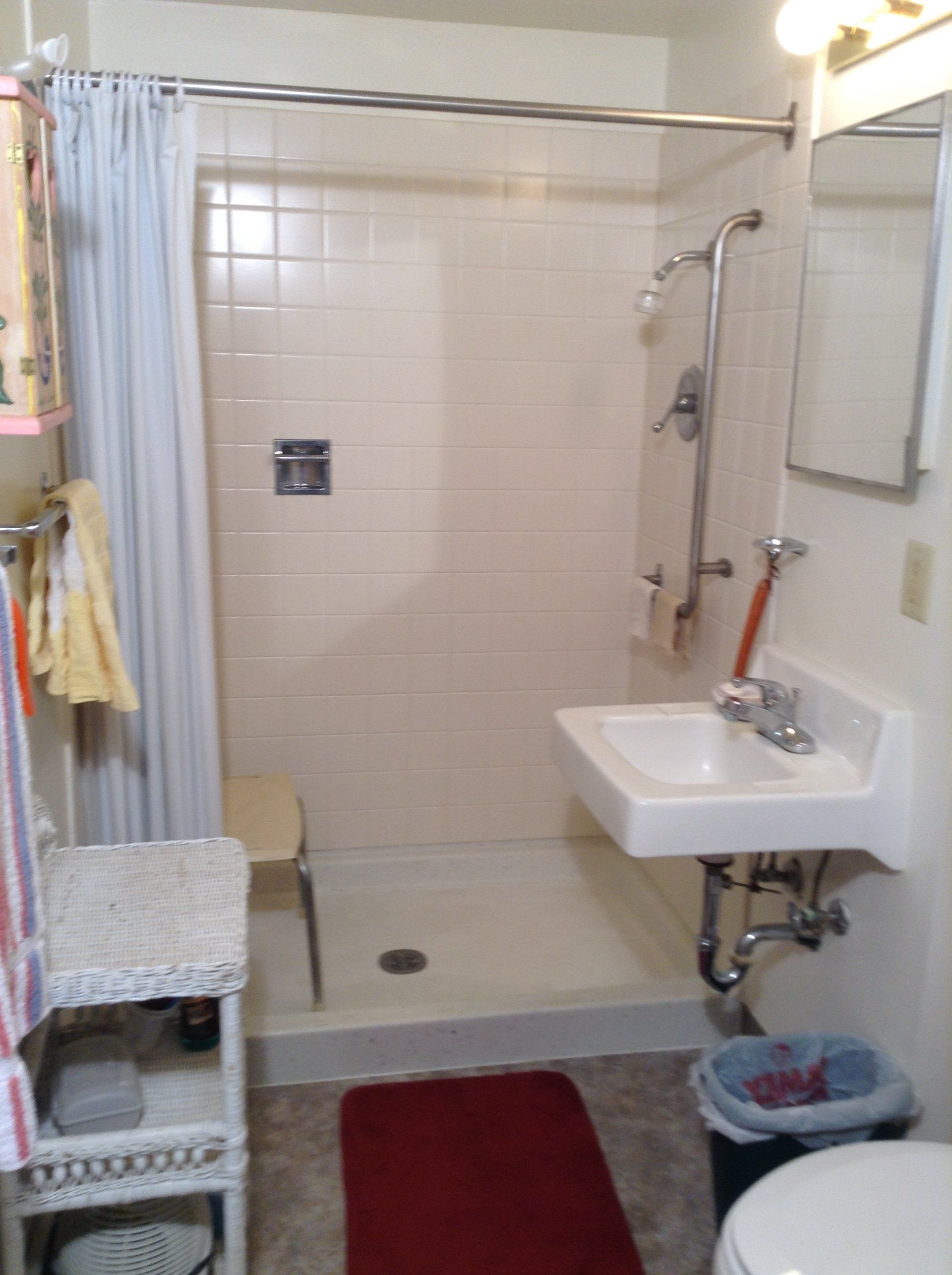 Small bathroom with a shower, sink, and toilet. White tile, red rug, and towels on a rack are visible.