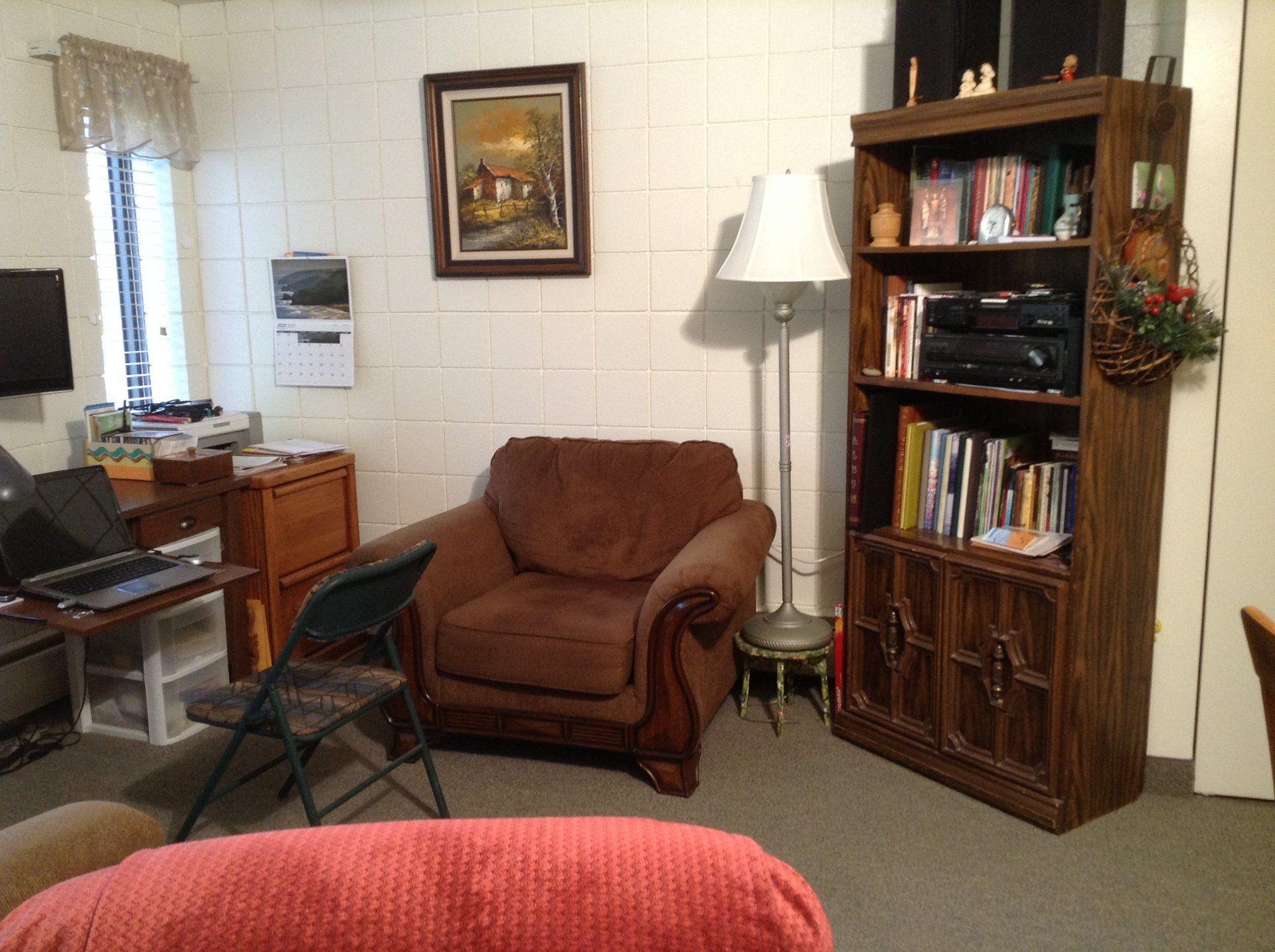Cozy living room with brown armchair, wooden bookshelf, lamp, and framed artwork on a brick wall.