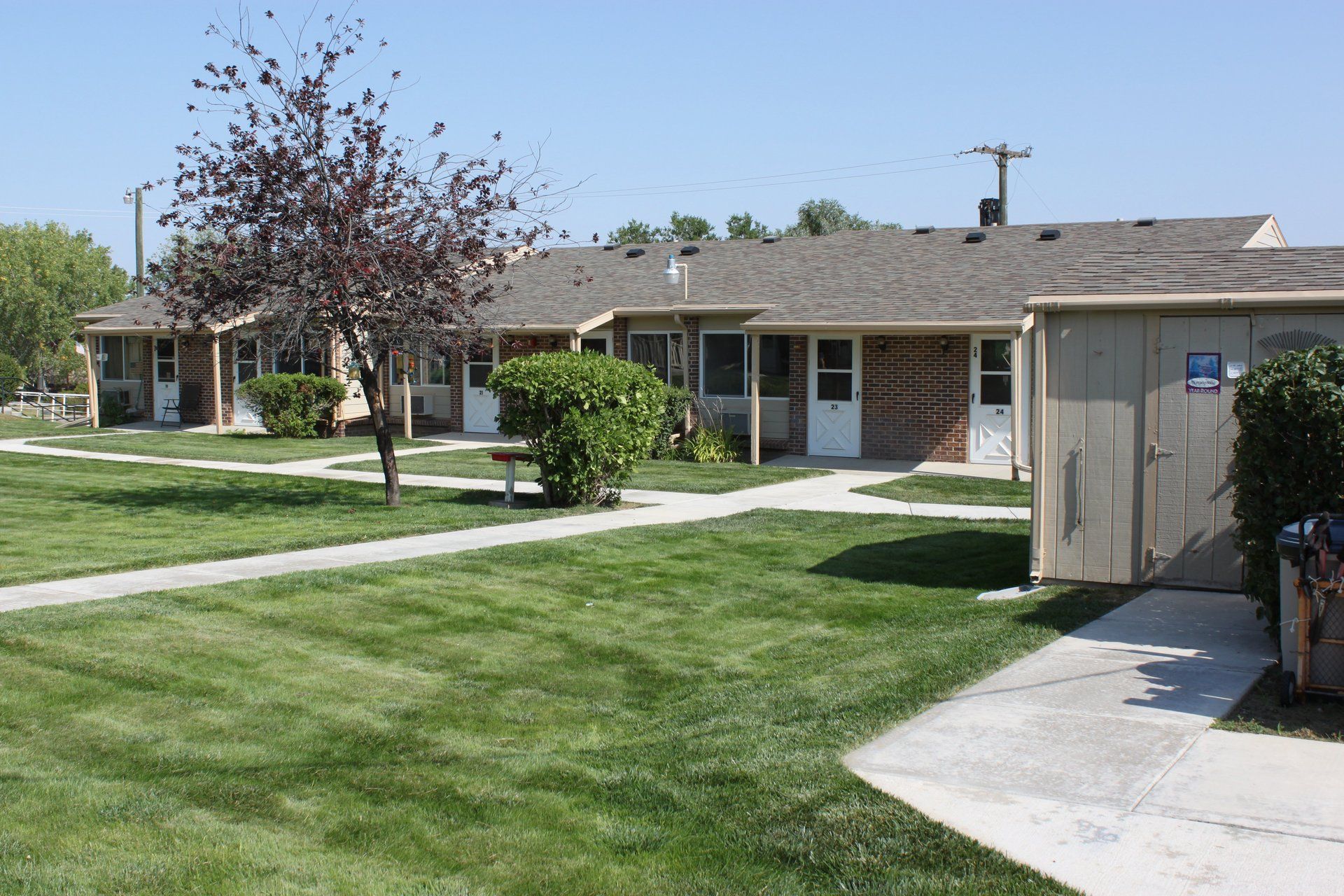 Row of low-rise brick buildings with green lawns and a clear sky. A tree and bushes are in the yard.