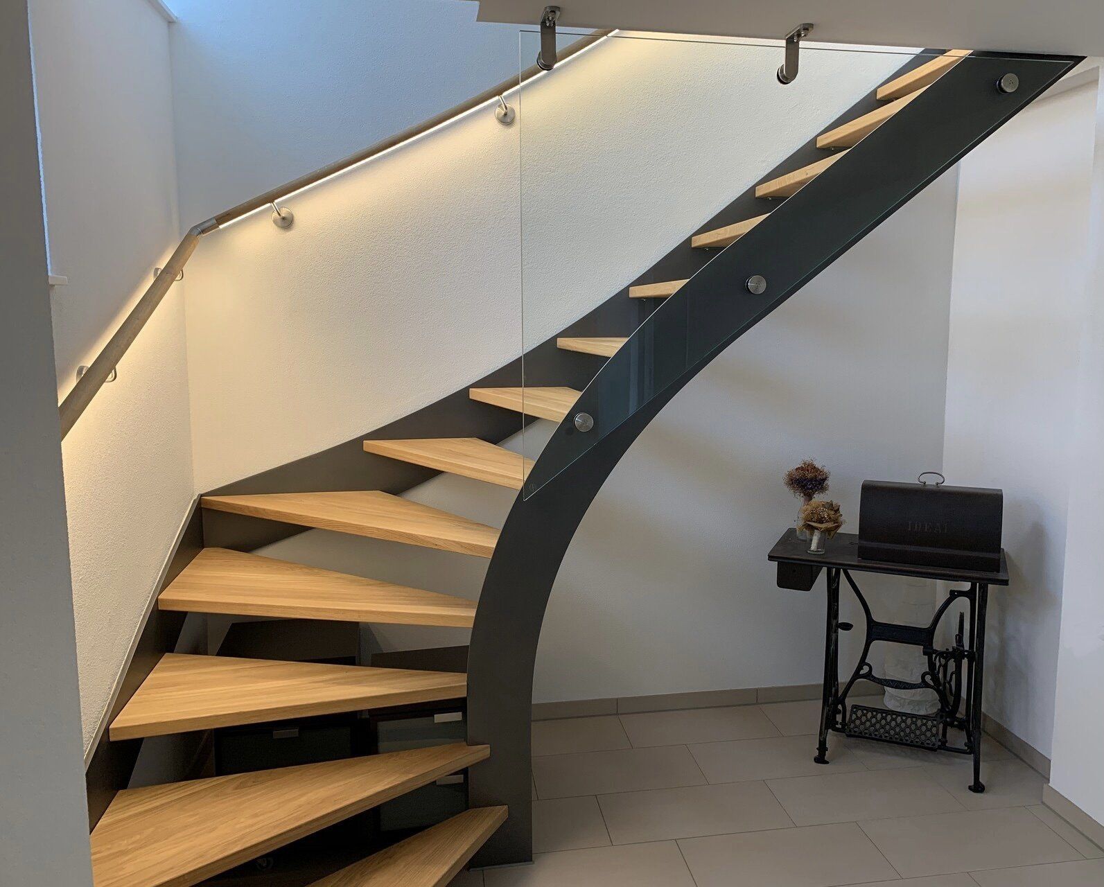 Eine hölzerne Wendeltreppe mit einem Glasgeländer in einem Raum neben einer Nähmaschine.