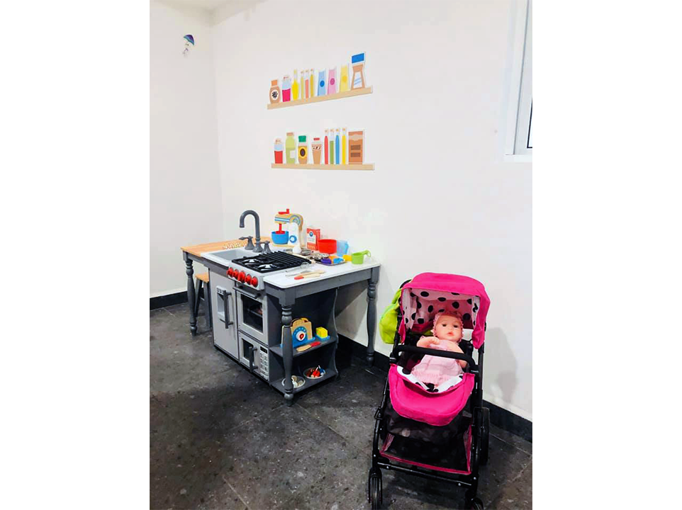 Una muñeca está sentada en un cochecito rosa junto a una cocina de juguete.