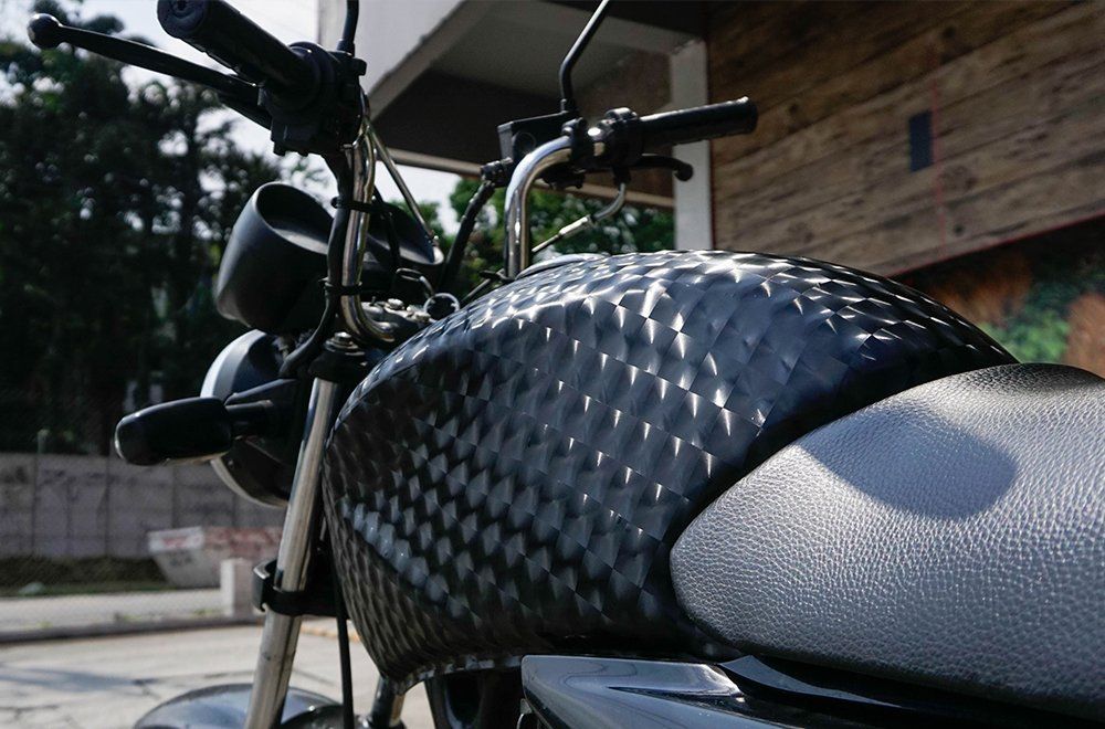 Detalhe do tanque de combustível de fibra de carbono preta com padrão texturizado de uma motocicleta, estacionada ao ar livre.